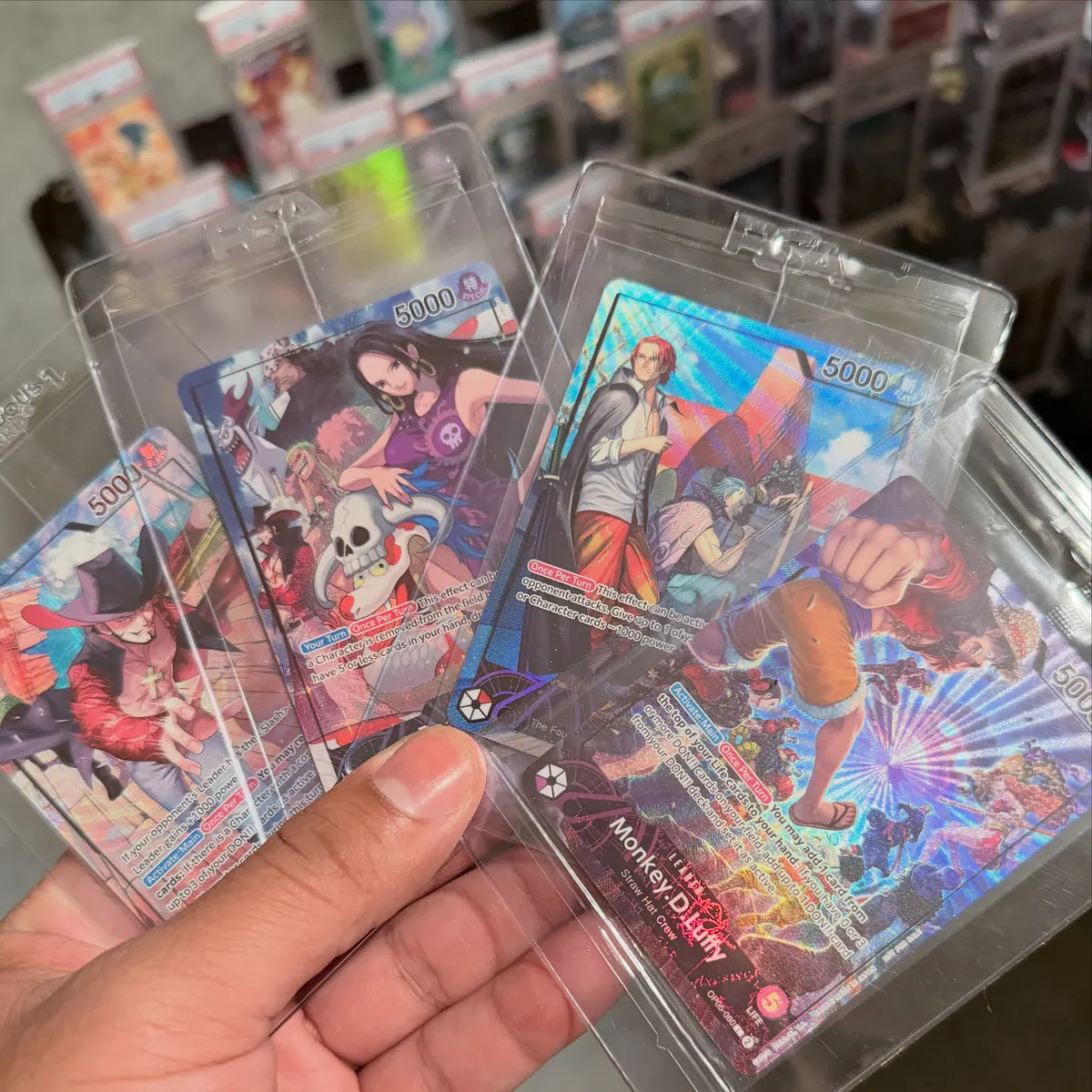 A hand fanning out four One Piece cards in top loaders, including Boa Hancock, Shanks and Monkey D. Luffy