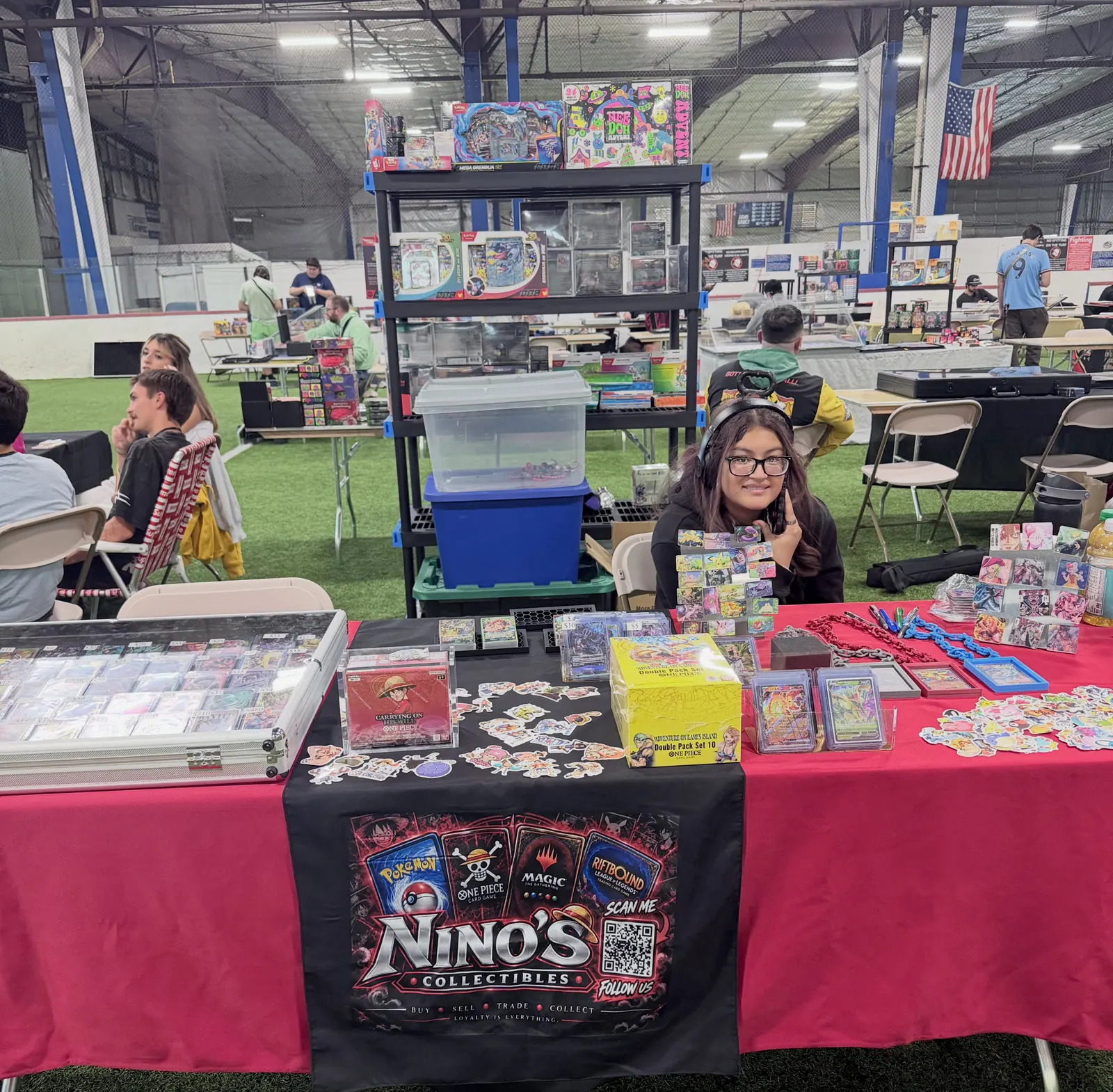The Nino's Collectibles vendor table at a card show: a red tablecloth, a black Nino's Collectibles banner, card cases and sealed product, with a helper seated behind it