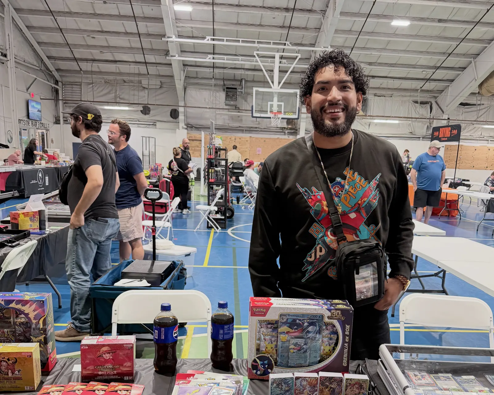 Nino smiling behind his vendor table at an indoor card show, with Pokémon and One Piece boxes and binders on the table