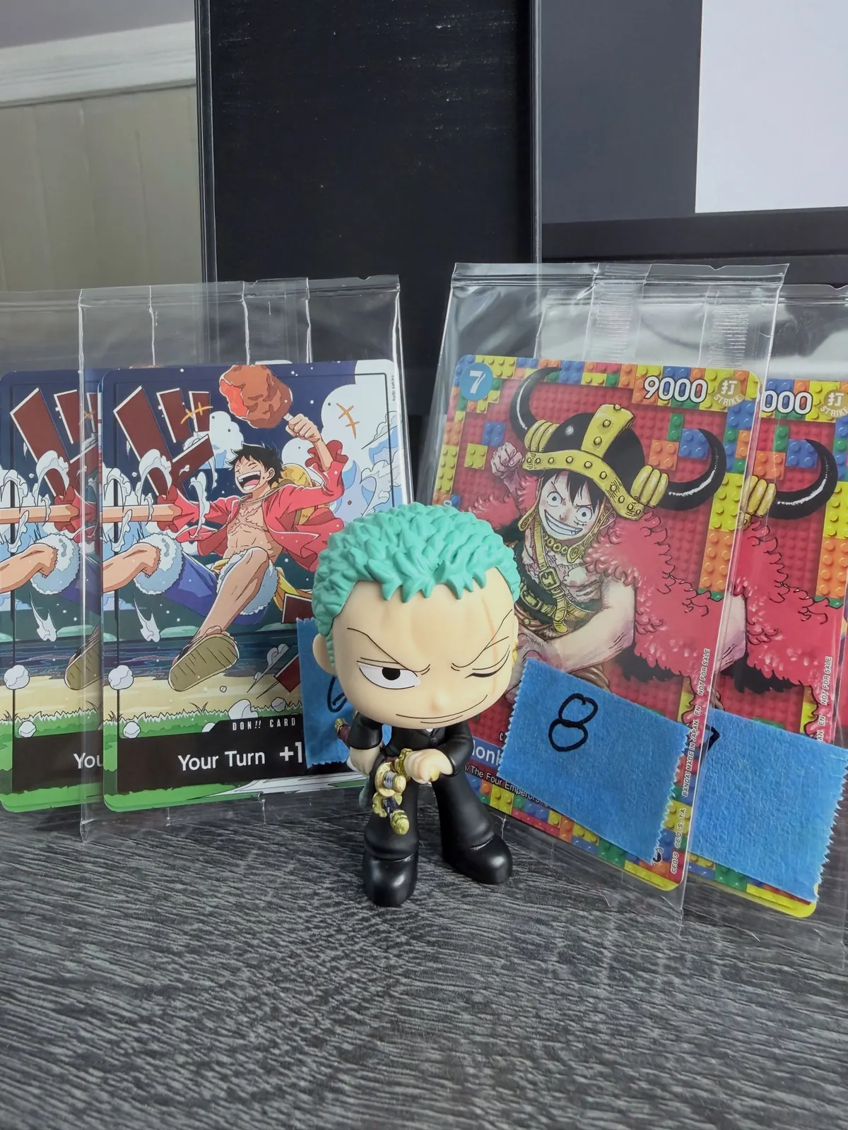 One Piece Luffy cards in sleeves lined up behind a Zoro figure on a desk