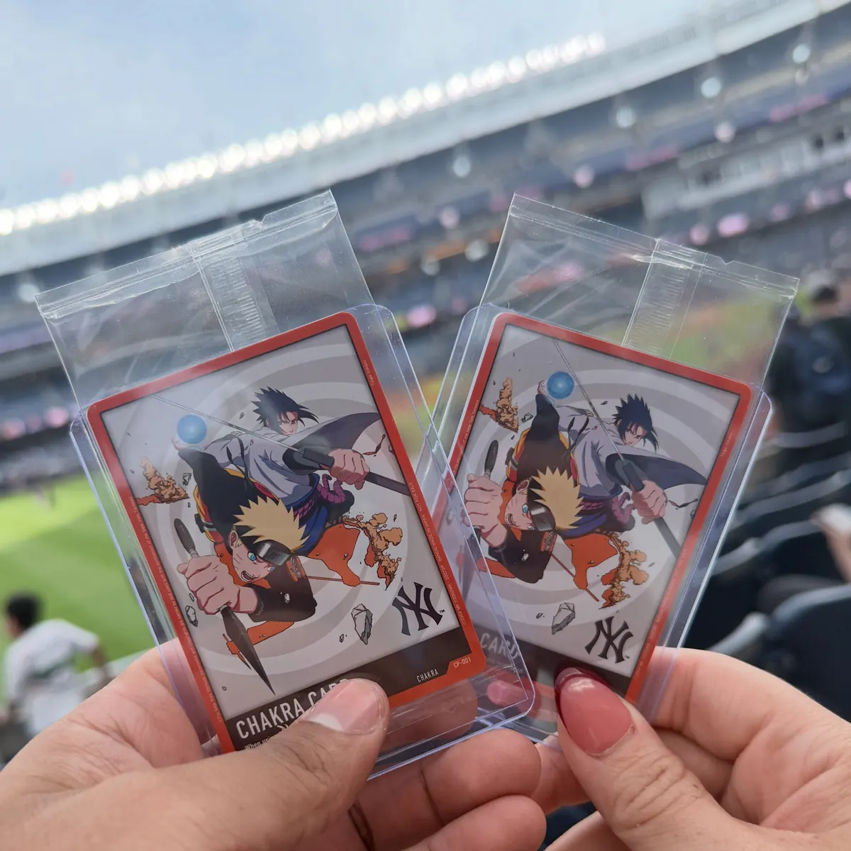 Two hands holding Naruto and Sasuke cards with Yankees logos, inside Yankee Stadium